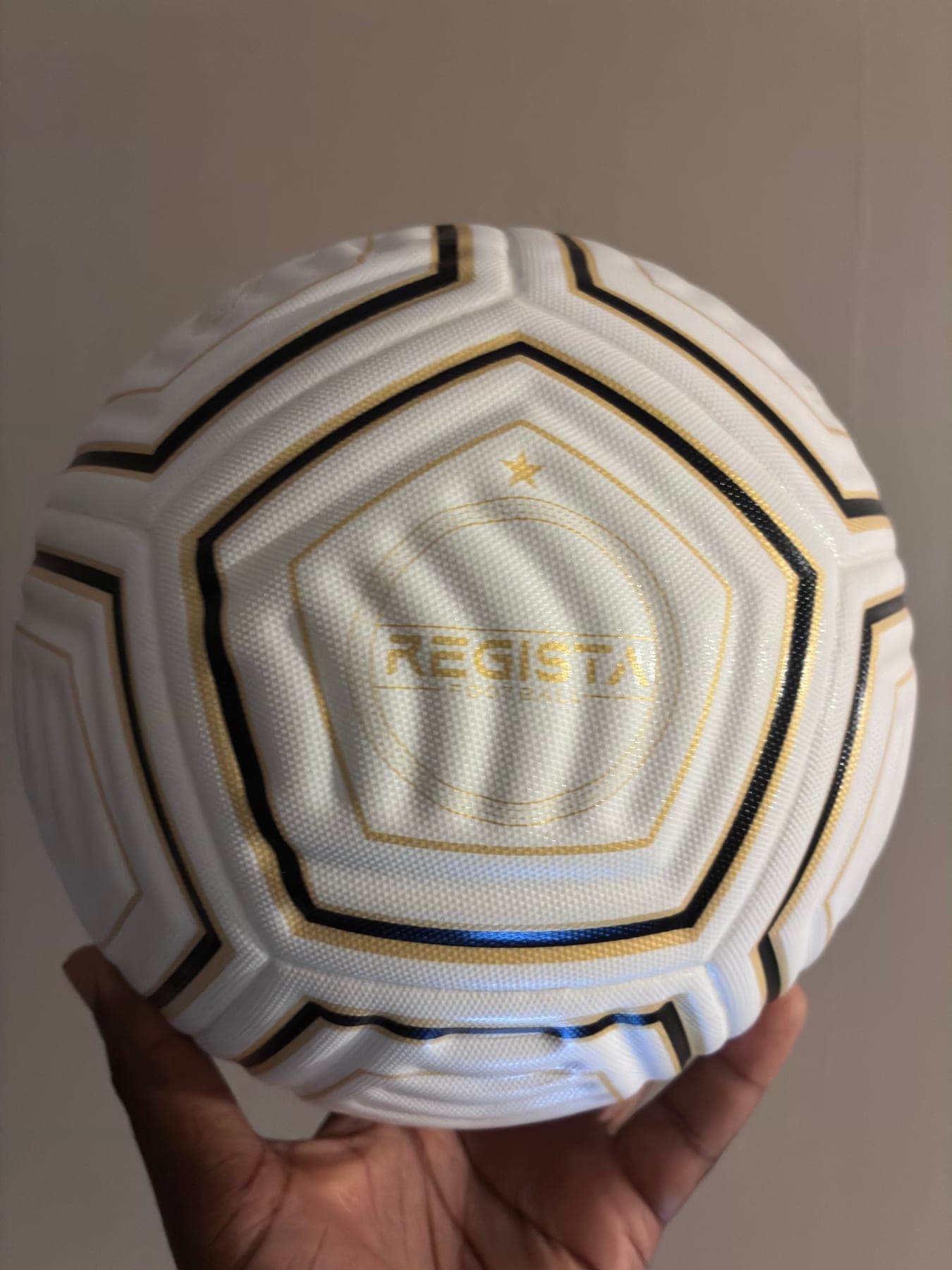 The Regista V1 Pro Match Ball, white with gold and black panel outlines and a Regista roundel on the front panel.